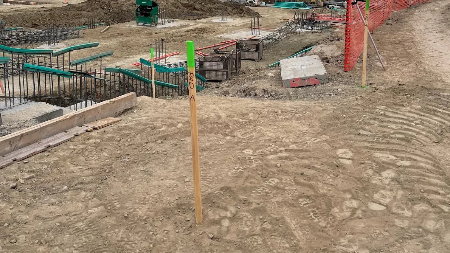 Commercial construction site with multiple concrete footings formed and poured, grade stake set, and a concrete truck working in the background
