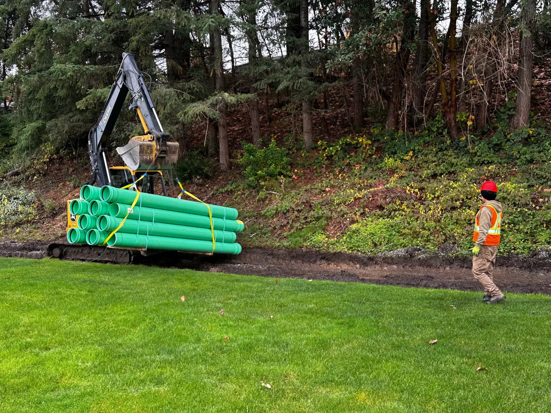 Bundle of green PVC storm drain pipe being delivered by excavator to a drainage trench on a treed site
