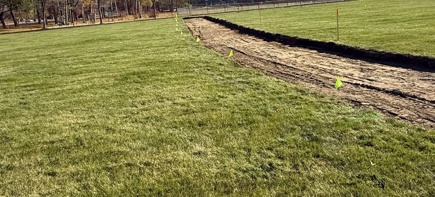 Graded drainage trench cut across a lawn with layout flags marking the drain line